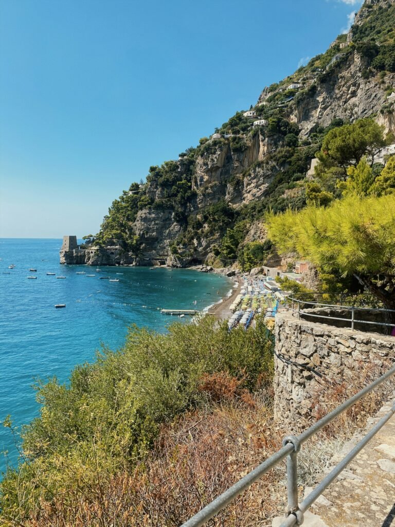 Scenic view of Positano beach with turquoise sea, sunbeds, and steep cliffs on Italy’s Amalfi Coast.