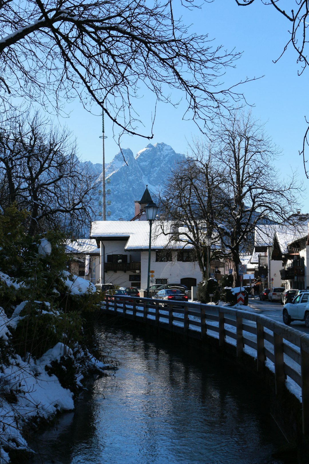 A snow-covered Bavarian-style village with a stream running past wooden footbridges and a jagged mountain peak rising behind the rooftops