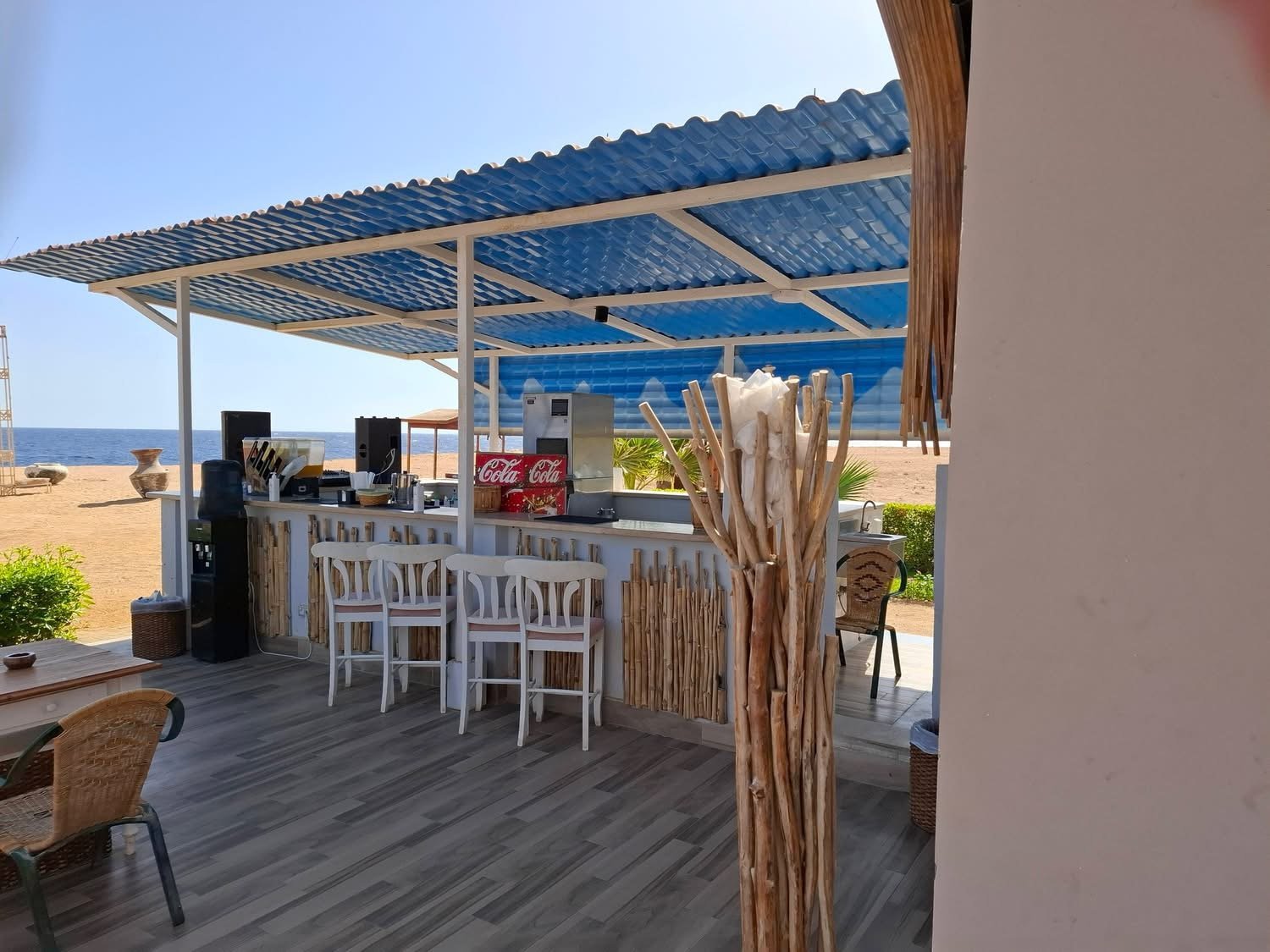 An open-air beach bar with a blue roof, white stools and a Coca-Cola fridge beside the sand and sea