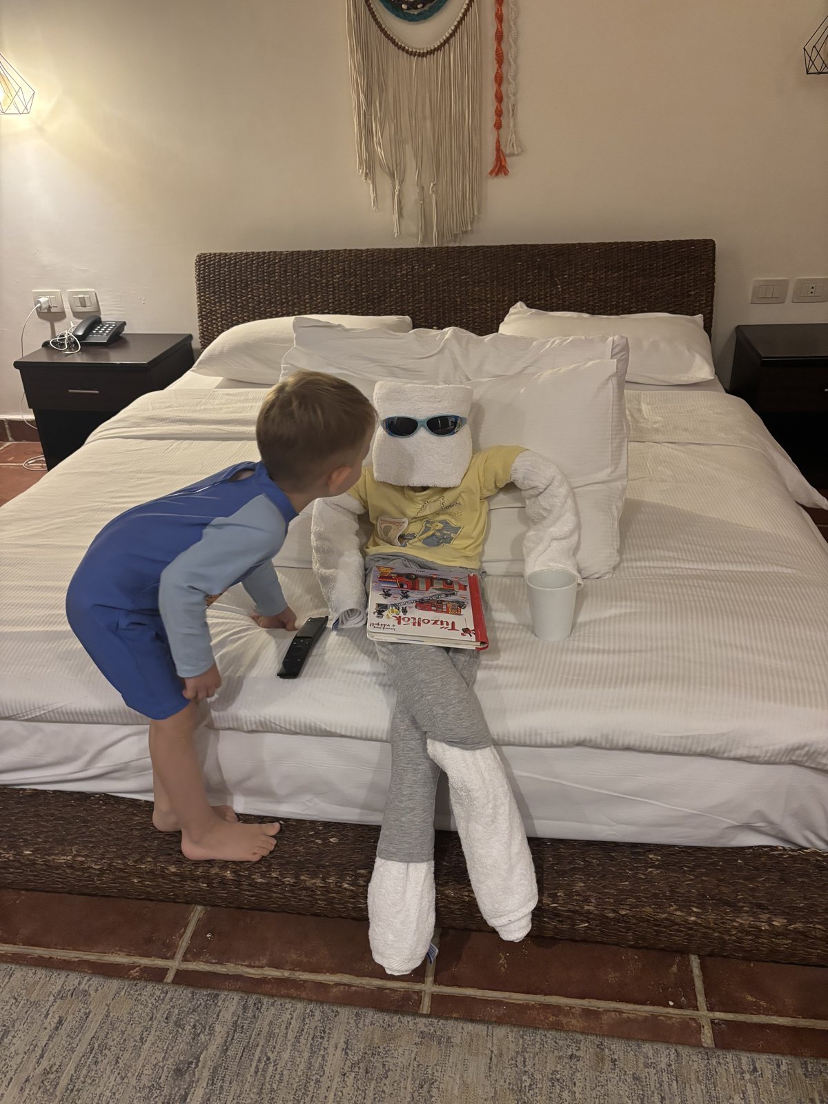 A hotel bedroom with a wicker headboard and white bedding, where a small boy plays with a towel figure wearing sunglasses