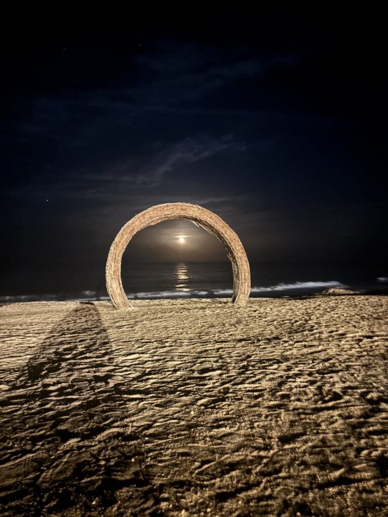 A giant wicker ring on a sandy beach at night with the moon shining through it