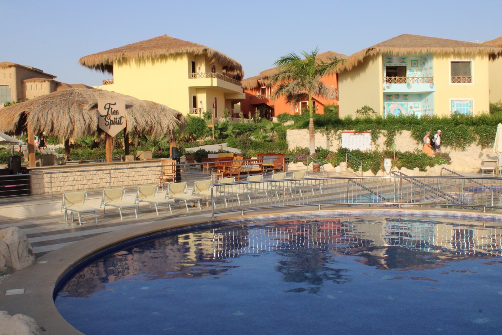 A curved resort pool with sun loungers in front of yellow thatched-roof guest houses and palm trees in bright daylight