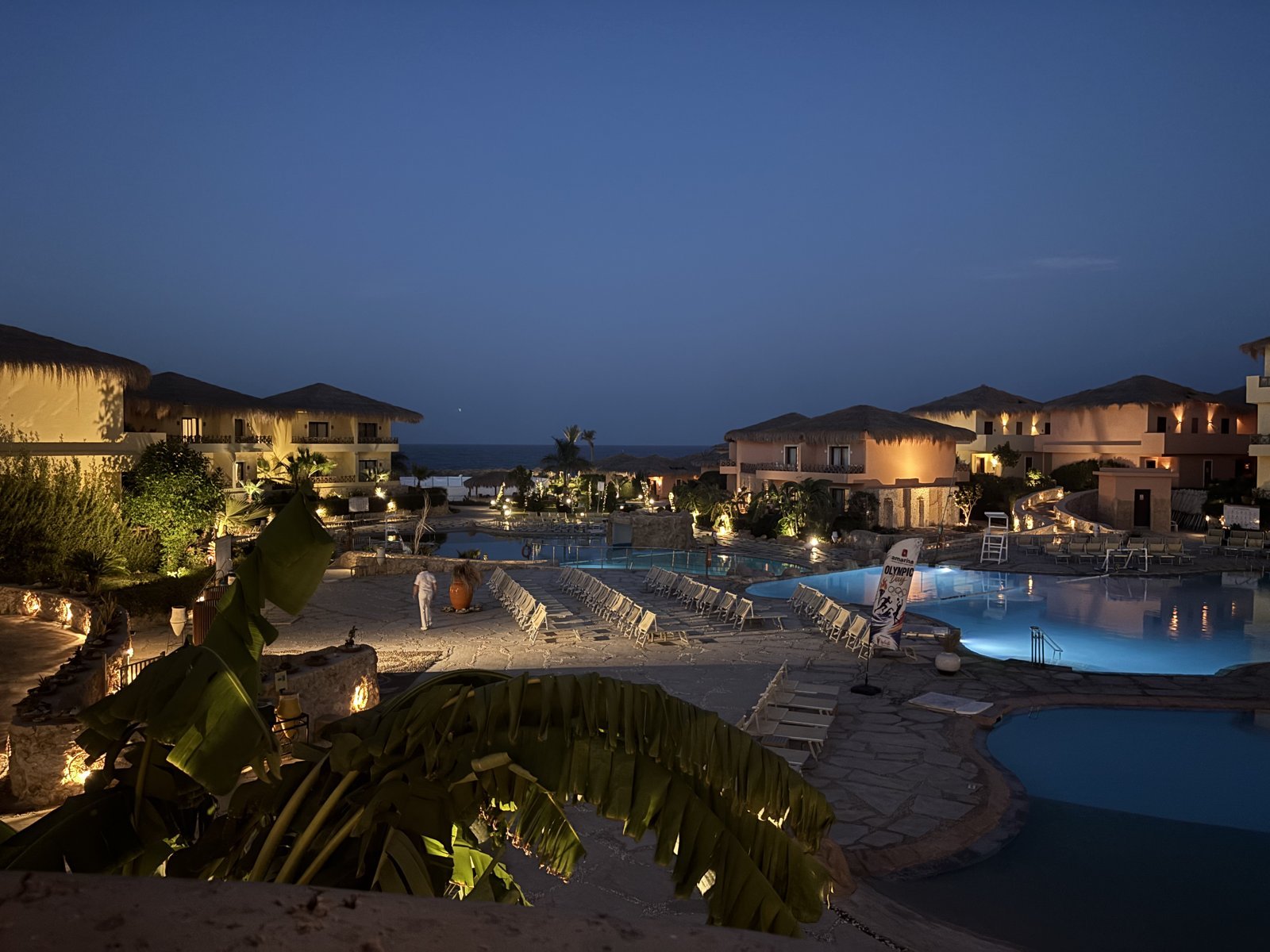 Resort pools and guest buildings lit up at dusk, seen from above through palm leaves