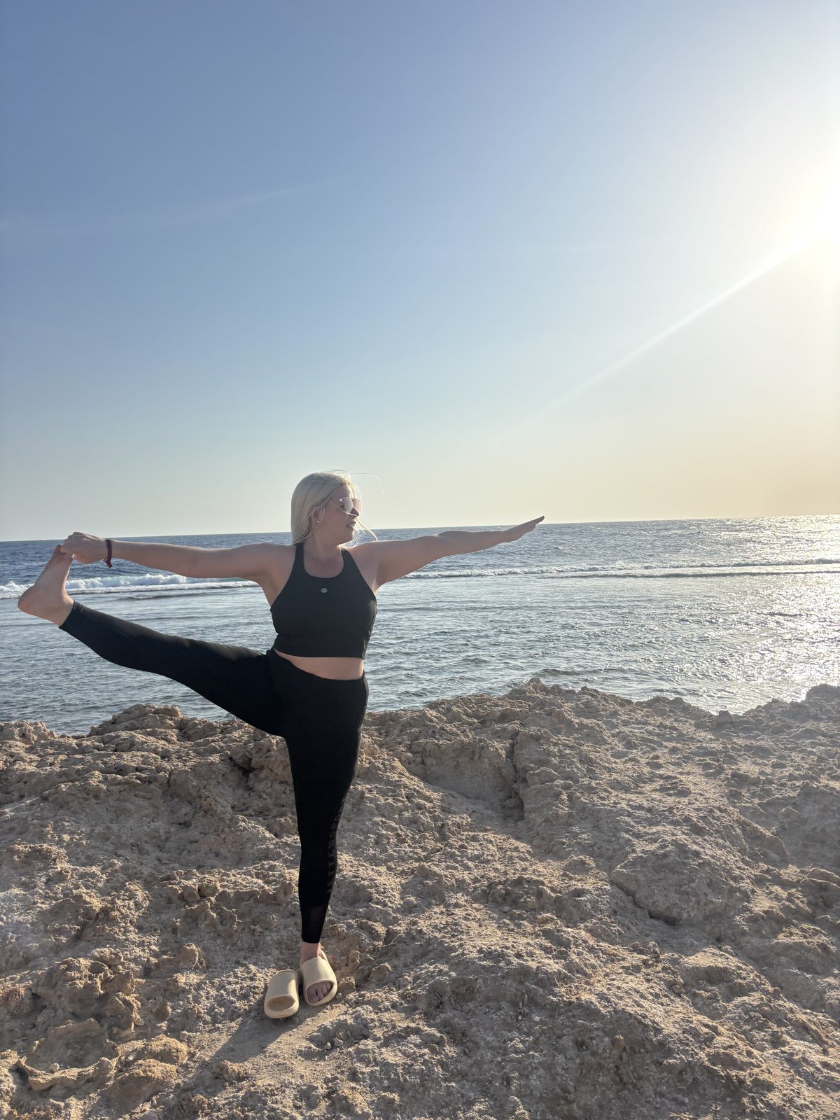 A woman holding a standing yoga pose on one leg on rocks by the sea in the morning sun