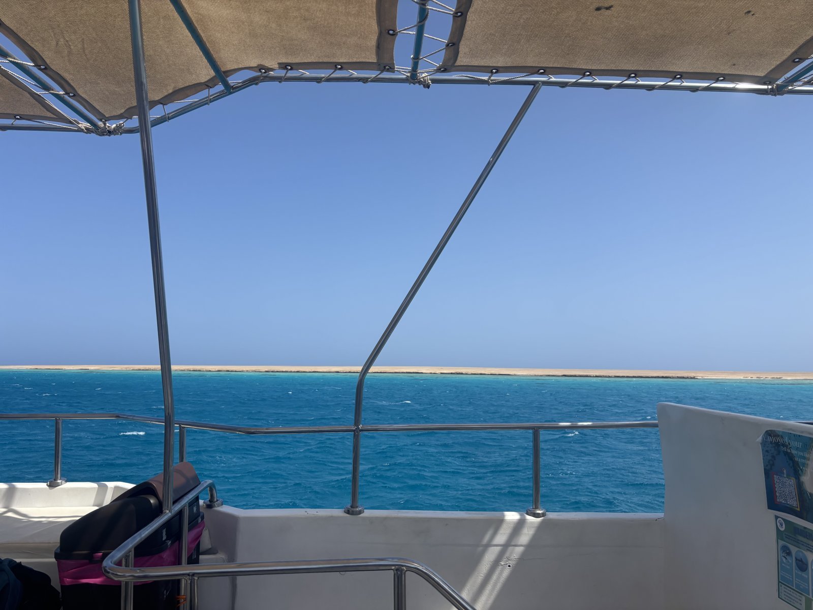 The deck of a day-trip boat under a canopy with the open Red Sea and a low sandy coast on the horizon